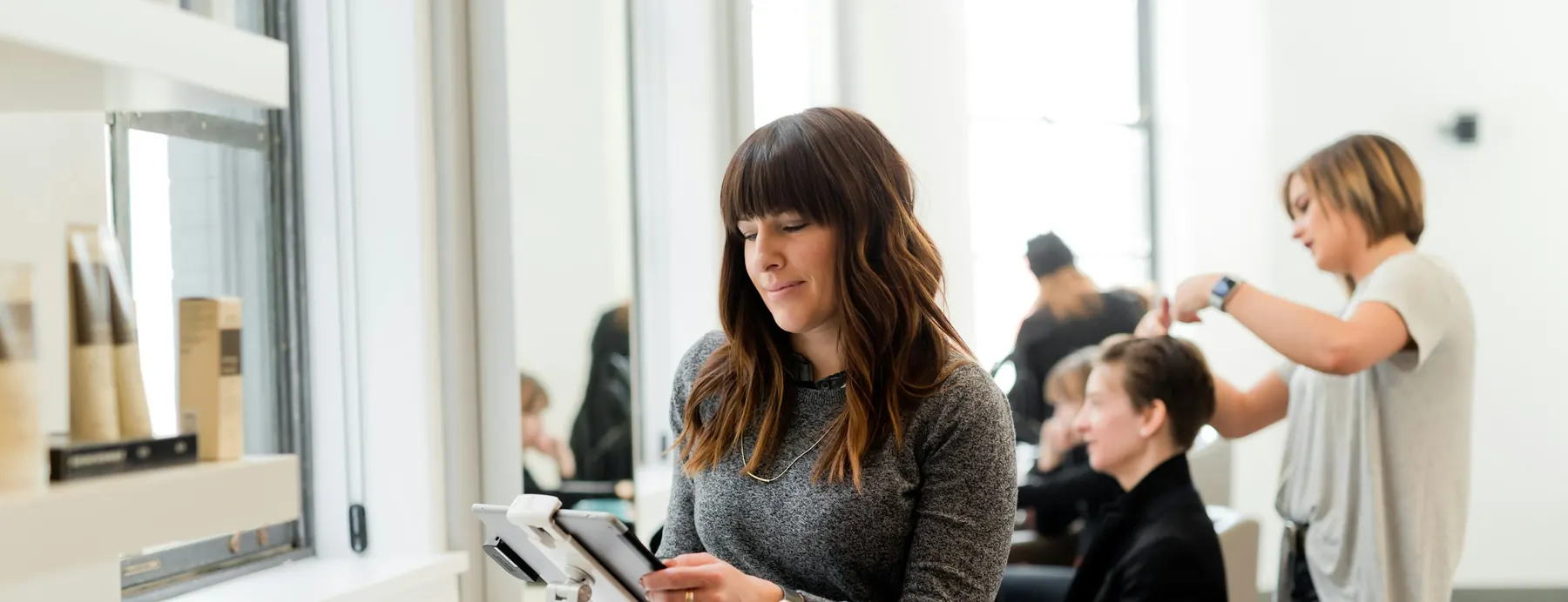 Coiffeuse qui consulte son planning sur tablette entre deux clientes, dans un salon lumineux